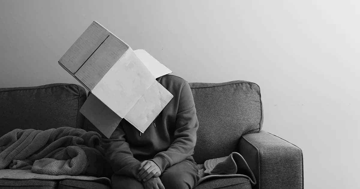 Persona sentada en un sofá con una caja de cartón en la cabeza, expresando el bloqueo emocional y la necesidad de evitar la realidad.