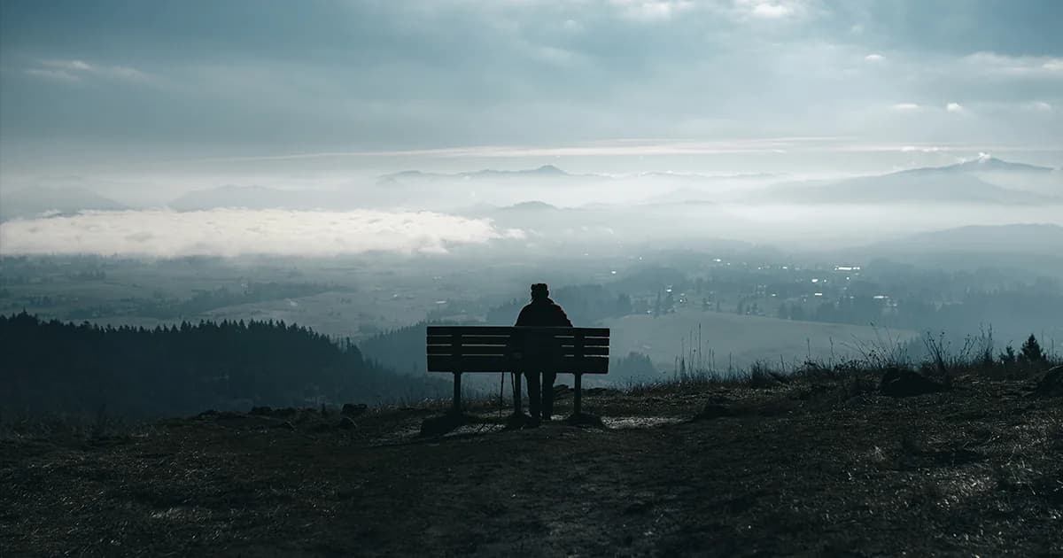 Persona sentada en un banco sobre la cima observando el valle con niebla, simbolizando una perspectiva amplia para encontrar dirección.